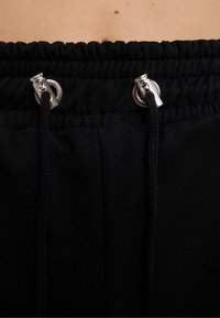 Černý pás joggerů se staženou elastickou částí, obsahující dvě stříbrné oka a splétanou šňůrku. Měkká, hladká textura.
