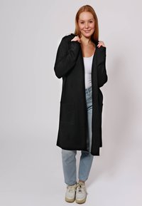Cardigan lungo nero con due tasche frontali, realizzato in un materiale morbido, indossato sopra una maglietta bianca e jeans azzurri, abbinato a sneakers bianche.