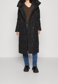 Kvinna iklädd en lång svart vadderad kappa, brun V-ringad tröja, blå jeans och svarta platåskor som står mot en enfärgad bakgrund.