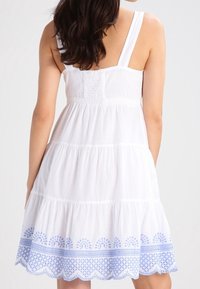 Vestido de sol blanco de algodón con un cuerpo ajustado, tirantes ajustables y una falda acampanada de volantes. Presenta bordados azules a lo largo del dobladillo.