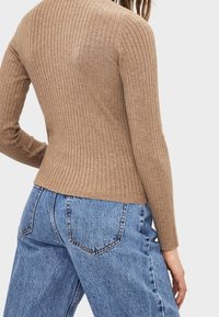Kvinna klädd i en åtsittande, ribbad, beige långärmad tröja, kombinerad med högmidjade, lösa blå denimjeans mot en enfärgad bakgrund.
