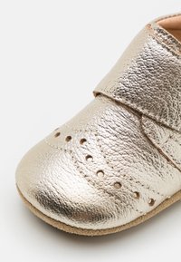 Metalická zlatá detská topánka s mäkkým koženým zvrškom, s velcro strapom a perforovanými detailami na špičke. Suede podrážka.