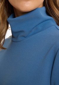 Nahaufnahme einer Person, die einen blauen Rollkragenpullover trägt, mit sichtbarem Hals und einem Teil des unteren Gesichts sowie braunem Haar.