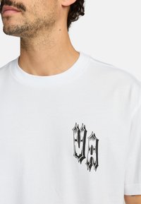 Witte katoenen t-shirt met een zwart grafisch ontwerp op de linkerkant van de borst. Klassiek ontwerp met ronde hals en korte mouwen. Zachte textuur.