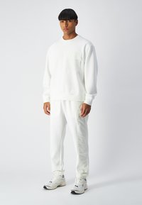 Sweatshirt blanc avec une poche plaquée, associé à un jogging blanc avec des panneaux latéraux texturés. Fini avec des baskets de couleur claire.
