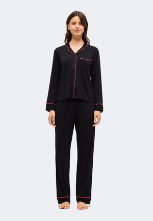 Mujer de pie descalza con pijama negro de manga larga con botones y ribete rosa en el cuello, puños, bolsillo y dobladillos del pantalón contra un fondo liso.