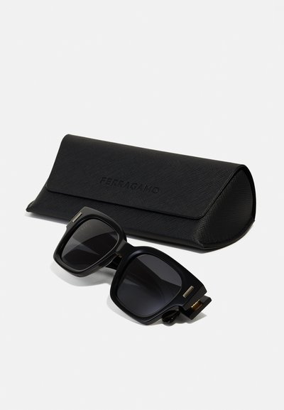 Occhiali da sole oversize neri con lenti scure, caratterizzati da una montatura robusta e dettagli in oro, esposti accanto a un astuccio nero texture con logo in rilievo.