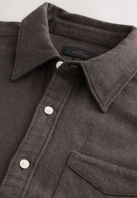Chemise en laine gris foncé à boutons, avec col pointu et poche poitrine, ornée de boutons blancs et d'une étiquette de taille moyenne.