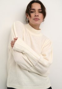 Pull en tricot couleur crème avec un col haut, des manches larges et des accents texturés. Il présente une coupe décontractée et une matière douce et légère.