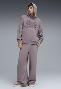 Sudadera lila de gran tamaño con diseño gráfico en el frente, combinada con pantalones anchos a juego. Ambos en tejido suave y texturizado. Zapatillas casuales completan el look.