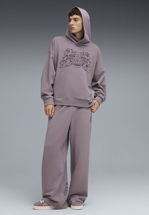 Przesadny fioletowy hoodie z grafiką na froncie, w zestawieniu z dopasowanymi szerokimi spodniami. Oba wykonane z miękkiego, textured materiału. Całość dopełniają casualowe sneakersy.