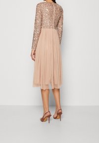 Abito in tulle beige con maniche lunghe, corpo impreziosito da paillettes, gonna plissettata e zip invisibile sul retro. Indossato con sandali con tacco color nude.