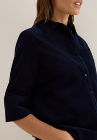 Donkerblauwe corduroy shirt met een knoopkraag, korte opgerolde mouwen en een borstzak. Gestructureerde stof met verticale lijnen.