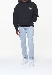 Hombre vistiendo una sudadera negra con un pequeño estampado blanco, jeans azul claro y zapatillas blancas, de pie contra un fondo blanco.