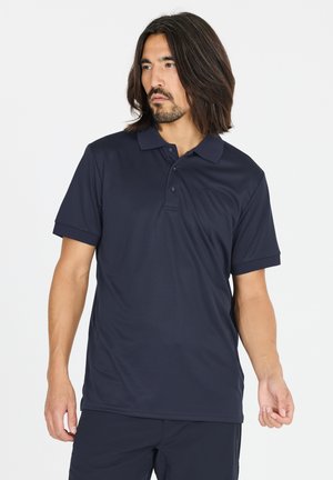 Mand med mellemlangt mørkt hår iført en marineblå kortærmet polo og matchende bukser, der ser mod venstre.