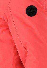 Koralfarvet jakke med glat tekstur, der har et sort cirkulært logo-patch og markante sømdetaljer på ærmet.