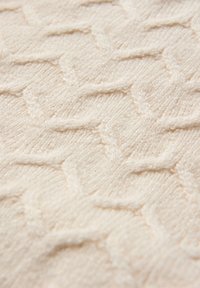 Kermanvärinen neulottu kangas, jossa on teksturoitu kuvio lomittain kiertyvistä siksak-silmukoista.