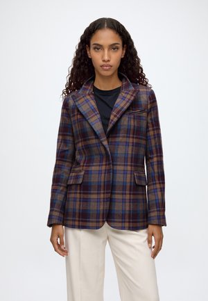 Una modelo posa sobre un fondo blanco liso, con una americana de cuadros marrones, azules y burdeos sobre un top negro y unos pantalones color crema.