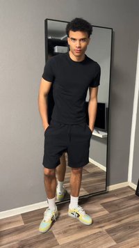 Νεαρός άντρας που φοράει μαύρο μπλουζάκι και σορτς στέκεται με τα χέρια στις τσέπες μπροστά σε έναν καθρέφτη ολόσωμο σε δωμάτιο με ξύλινο πάτωμα.