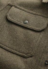 Olive green wool blend jacket with a front pocket and a silver snap button. Soft texture and clean lines with a classic design.
