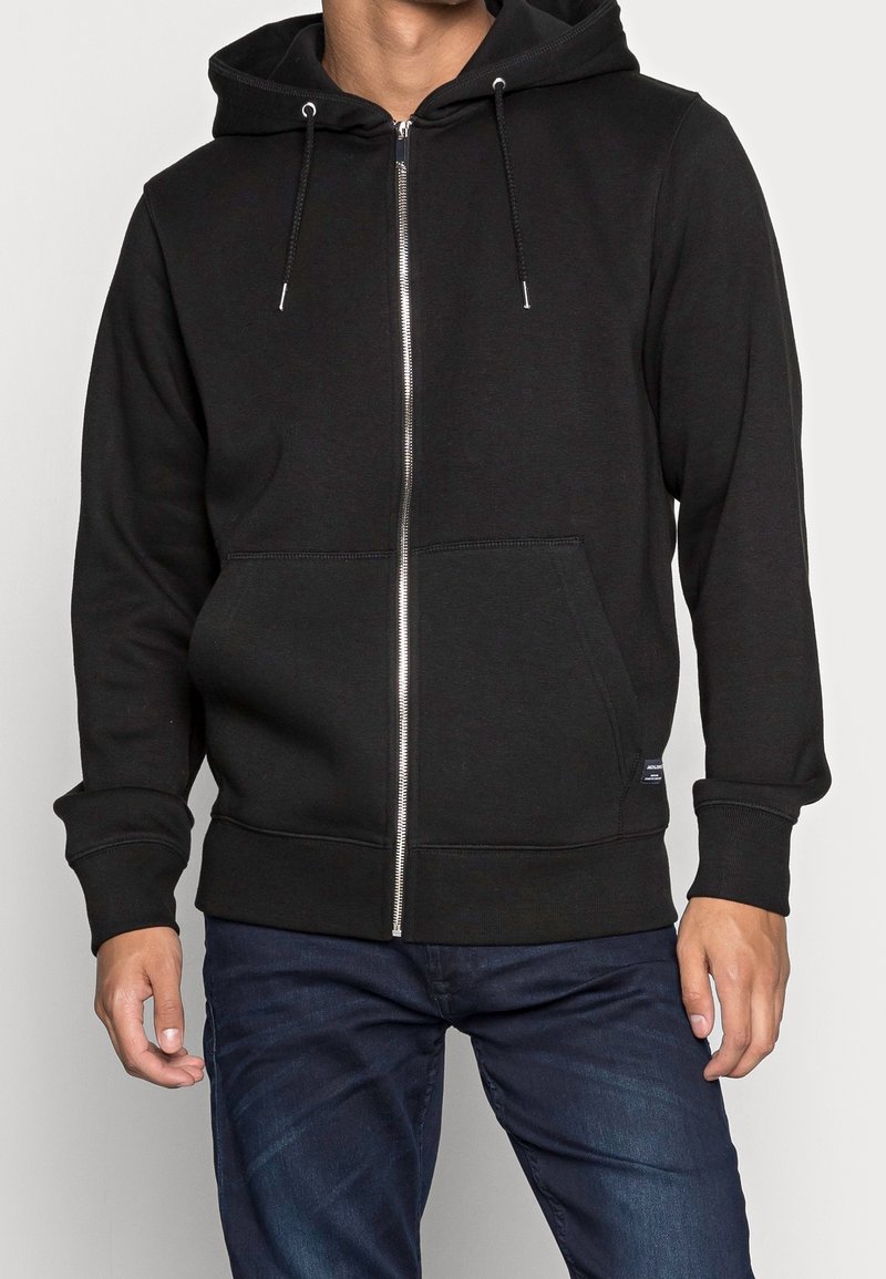Homme portant un sweat à capuche noir zippé avec poches avant et un jean bleu, debout devant un fond clair uni.