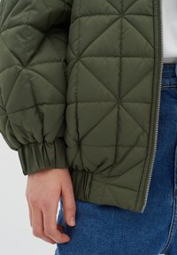 Olivově zelená prošívaná bunda s diamantovým vzorem, s elastickými manžetami a stříbrným zipem; oblečená přes modré džíny s viditelnou rukou.