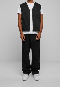 Czarna bezrękawnik z dekoltem w kształcie V, z dwiema kieszeniami z przodu, w połączeniu z luźnymi czarnymi spodniami i białymi sneakersami.