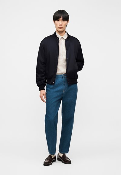 Junger Mann trägt eine schwarze Bomberjacke, ein beiges Hemd mit Knöpfen, blaue Jeans und dunkelbraune Loafers und steht vor einem schlichten weißen Hintergrund.