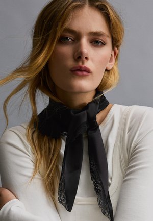 Blondynka o niebieskich oczach, ubrana w białą bluzkę i czarny szalik z koronkowymi wykończeniami związany wokół szyi na szarym tle.