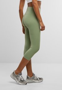 Groene, hooggetailleerde leggings met een gladde textuur, cropped lengte en platte naden, gecombineerd met grijze sportschoenen met witte veters.