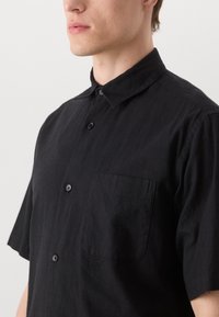 Camicia nera a maniche corte realizzata in tessuto strutturato, con colletto classico, pattina con bottoni frontale e una singola tasca sul petto.