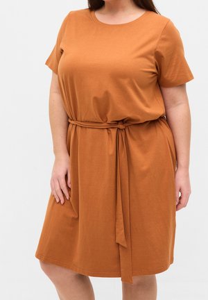 Femme portant une robe rouille à manches courtes, longueur genoux, avec une ceinture en tissu nouée à la taille, debout devant un fond clair uni.