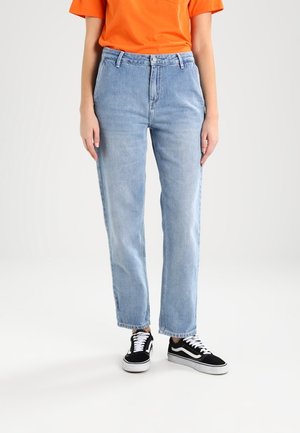 Person iført lyseblå jeans med lige ben, orange kortærmet skjorte og sort-hvide sneakers, stående på hvid baggrund.