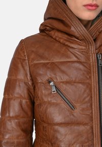 Chaqueta de cuero marrón con un diseño acolchado, capucha y bolsillo con cremallera. Presenta una textura suave y adornos de hardware plateados.