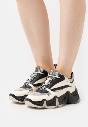 Baskets épaisses blanches et noires avec détails en mesh et cuir portées aux pieds sur un fond blanc uni.