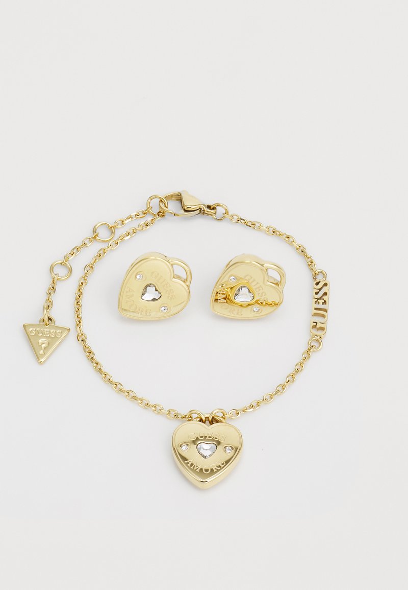Ensemble de bijoux en or comprenant un bracelet en forme de cœur avec un fermoir et un charm, ainsi que des boucles d'oreilles en forme de cœur assorties ornées de strass.