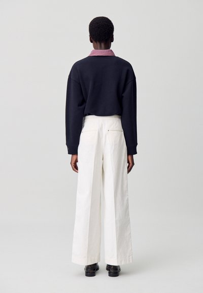 Sudadera negra con un cuello amplio sobre una camisa de cuadros rojos, combinada con pantalones blancos de pierna ancha y zapatos negros.