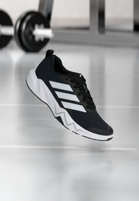 Black and white Adidas athletic shoe suspended above a reflective floor with a blurred barbell in the background.