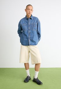 Denim ing, világoskék, két mellzsebbel; krémszínű sorttal párosítva; fekete slip-on cipőkkel; fehér zoknikkal viselve zöld padlón.