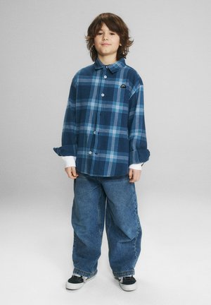 Chłopiec o brązowych włosach do ramion, ubrany w niebieską koszulę w kratę, luźne niebieskie dżinsy i czarno-białe trampki, stojący na szarym tle.