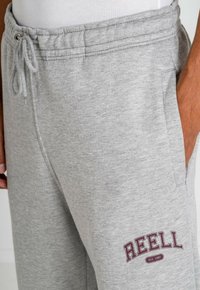 Graue sweatpants aus weichem Material mit elastischem Bund, Kordelzug, Seitentaschen und einem burgunderfarbenen "REELL"-Logo auf dem Oberschenkel.