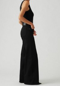 Femme portant un haut sans manches noir et un jean large taille haute noir, se tenant de profil avec une main dans la poche.