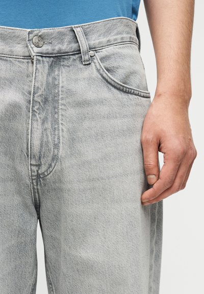 Κοντινό πλάνο στη μέση και πλαϊνή τσέπη τζιν άνοιχτου γκρι χρώματος, με το δεξί χέρι ενός ατόμου να ακουμπάει δίπλα στο ισχίο.