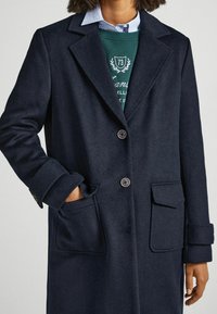Modello che indossa un cappotto blu scuro con due tasche frontali, sovrapposto a un felpa verde e una camicia azzurro chiaro, mostrando uno stile casual.