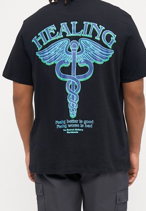 T-shirt noir avec symbole caducée bleu et vert, texte "HEALING" au-dessus, et phrases "Se sentir mieux est bon, se sentir pire est mauvais" sous le symbole.