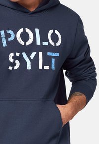 Marinblå huvtröja med en framficka, prydd med en slitagen tryck av "POLO SYLT" i vitt och ljusblått. Blandning av bomull, avslappnad design.
