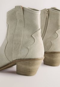 Lichtgroene suède enkellaarzen met een gestapelde hak, voorzien van golvende stikpatronen en een ritssluiting aan de zijkant.