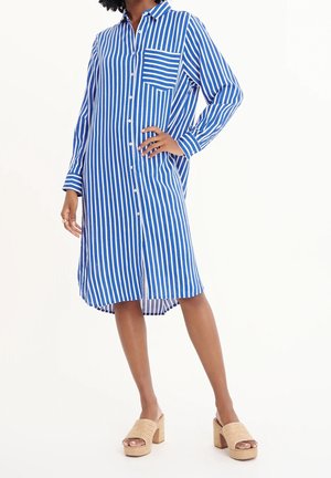 Frau, die ein blau-weiß vertikal gestreiftes Hemdkleid mit langen Ärmeln und beige Plateausandalen trägt.