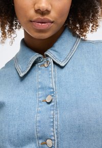 Άτομο με σγουρά μαλλιά που φοράει ένα ανοιχτό μπλε denim μπουφάν με κουμπωμένο το γιακά, με κοντινή λήψη στα χείλη και το κάτω μέρος του προσώπου.