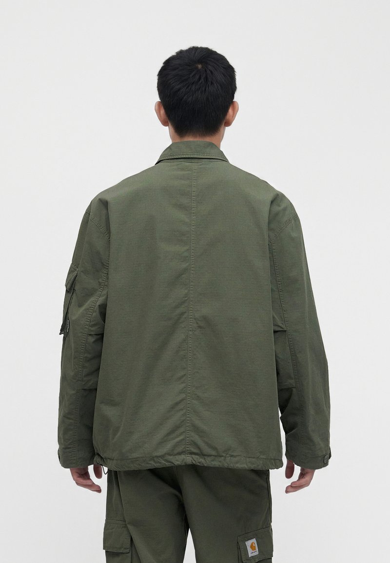 Hombre con chaqueta verde oliva y pantalones cargo, de pie de espaldas sobre un fondo gris claro liso.